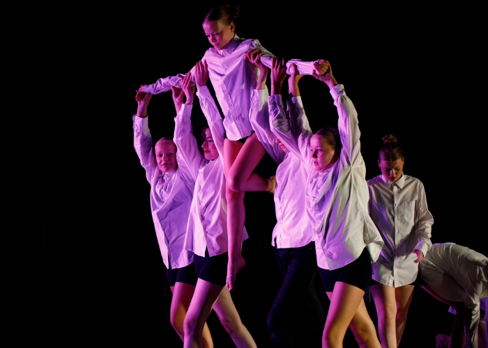 Groep dansers in witte overhemden lift een vrouw hoog in de lucht onder paarse verlichting.