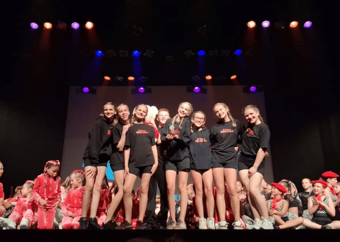 Een groep jonge dansers in zwarte shirts staat op een podium met een trofee, omringd door andere dansers in rode kostuums